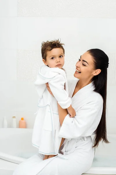 Mutter lacht und schaut Kleinkind-Sohn im weißen Bademantel an — Stockfoto