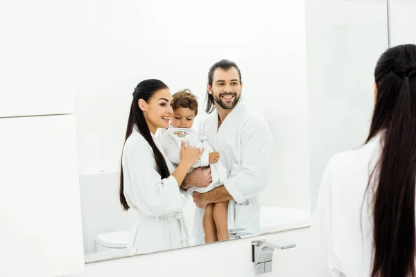 Mamá y papá mirándose en el espejo y sosteniendo a su hijo pequeño con batas blancas - foto de stock