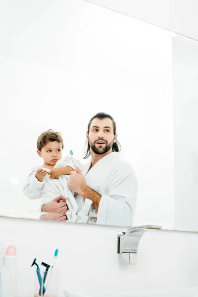 Pai e filho olhando no espelho em roupões de banho brancos e segurando escovas de dentes — Fotografia de Stock