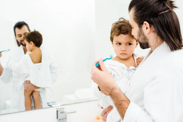 Papá mirando a su hijo y mostrando cepillo de dientes con pasta de dientes en el baño blanco - foto de stock