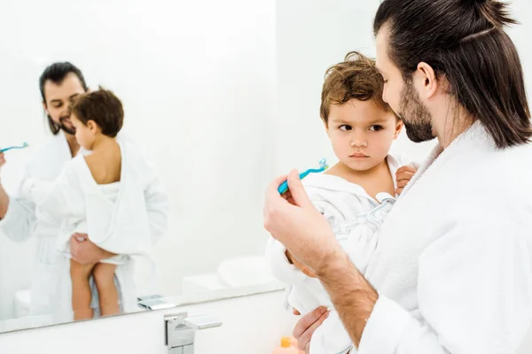 Dad looking at son and showing tothbrush with toothpaste in white bathroom — Stock Photo