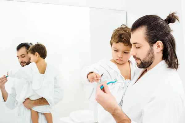 Hijo pequeño mirando el cepillo de dientes con pasta de dientes y papá sosteniéndolo en el baño blanco - foto de stock