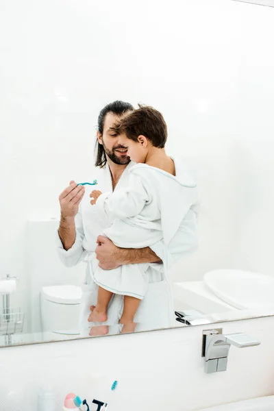 Père tenant tout-petit fils et montrant brosse à dents avec dentifrice dans la salle de bain blanche — Photo de stock