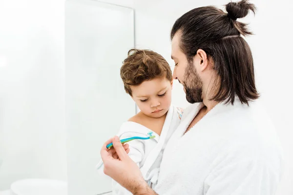 Papá mostrando cepillo de dientes con pasta de dientes a su hijo pequeño - foto de stock