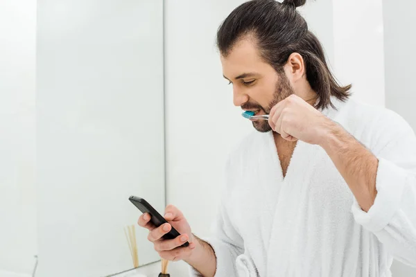 Hombre adulto guapo usando teléfono inteligente y cepillarse los dientes en el baño - foto de stock