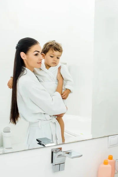 Beautiful mother holding son in white bathrobe in bathroom — Stock Photo