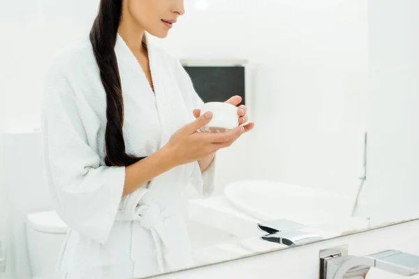 Vista recortada de la mujer en blanco albornoz celebración de la crema corporal - foto de stock