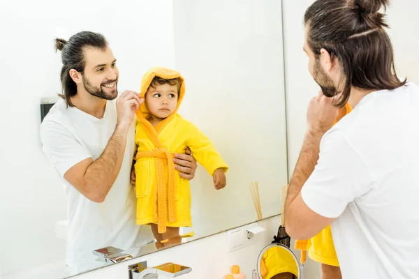 Sonriente padre celebración lindo niño hijo en amarillo albornoz en cuarto de baño - foto de stock