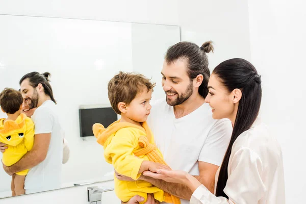 Feliz padres celebración lindo niño hijo en amarillo albornoz en cuarto de baño - foto de stock