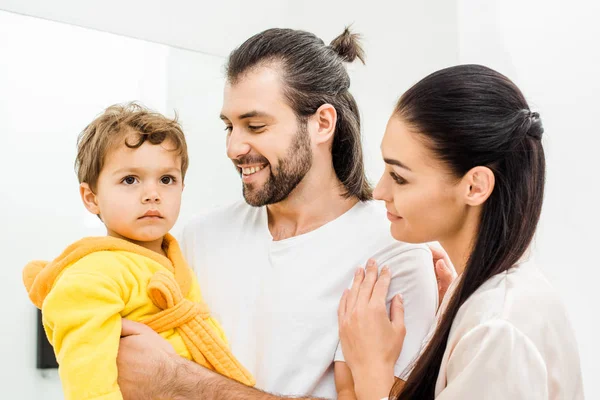 Улыбающиеся родители держат милого малыша сына в желтом халате в ванной комнате — стоковое фото