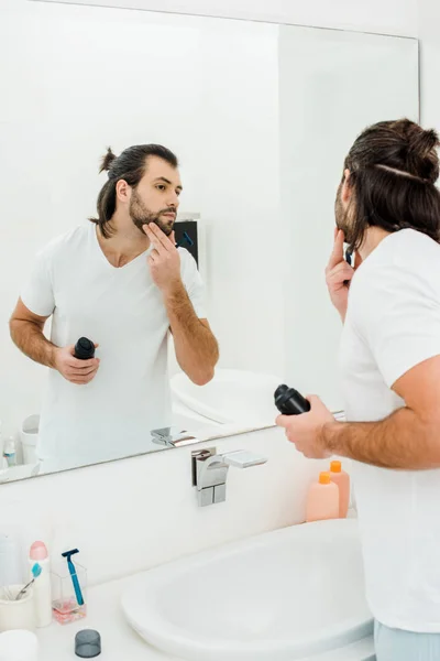 Bell'uomo che tiene schiuma da barba e si guarda allo specchio in bagno — Foto stock