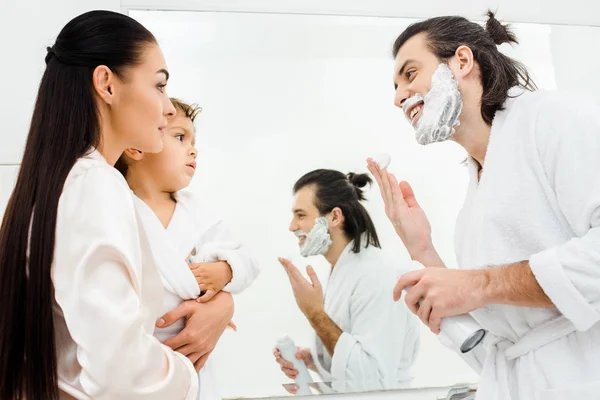 Sonriente hombre con espuma en la cara mirando a la esposa y el hijo en el baño - foto de stock