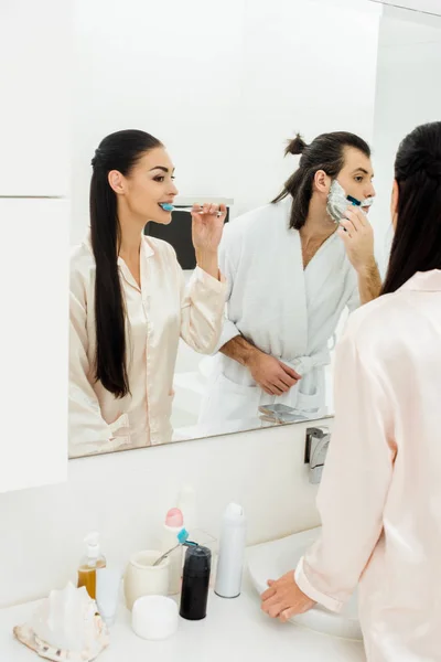 Bella donna lavarsi i denti e bello uomo rasatura davanti allo specchio in bagno — Foto stock