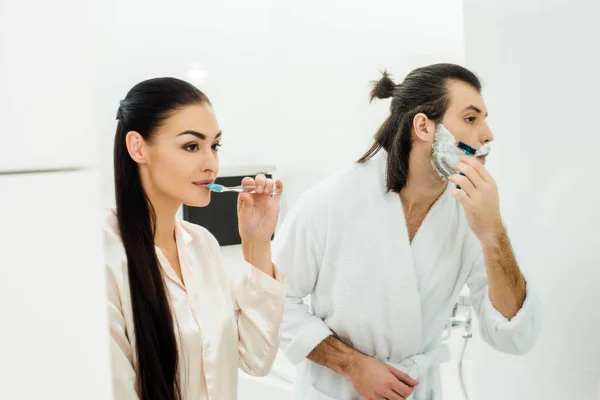 Hermosa mujer limpiando dientes mientras marido afeitando barba - foto de stock