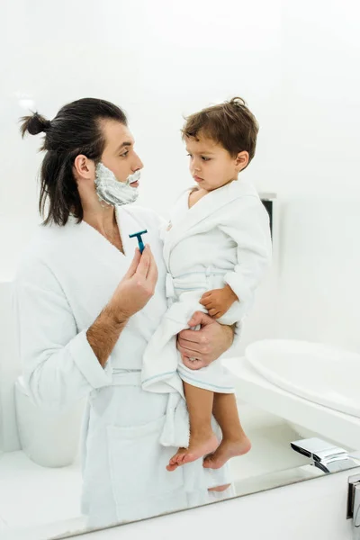 Vater im weißen Bademantel zeigt Rasiermesser auf Kleinkind-Sohn — Stockfoto