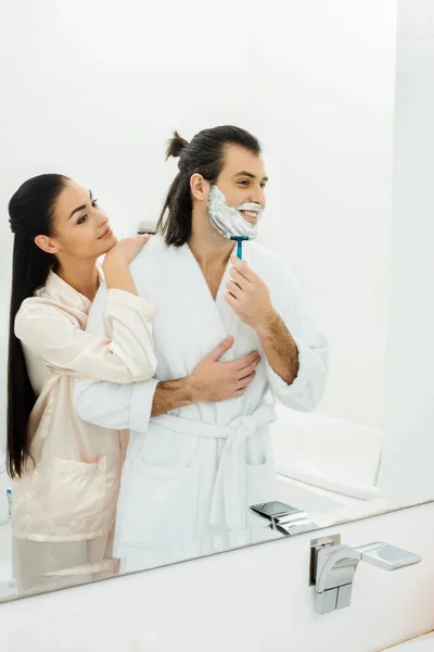 Mulher bonita abraçando marido enquanto homem de barbear na frente do espelho — Fotografia de Stock