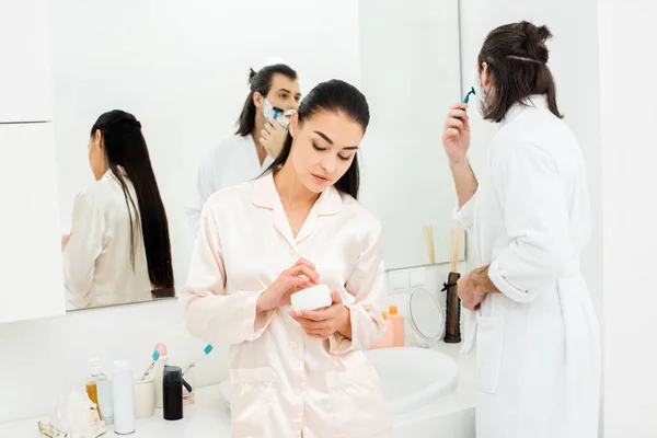 Hübsche Frau bleibt vor dem Spiegel, während Mann sich im weißen Badezimmer rasiert — Stockfoto