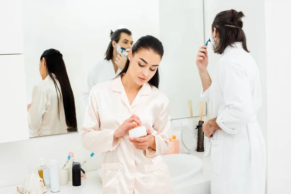 Mujer bonita que se queda delante del espejo mientras que el hombre afeitándose en el baño blanco - foto de stock