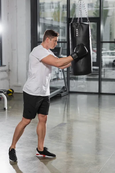 Vista lateral do treinamento bonito boxeador muscular com saco de perfuração no ginásio — Fotografia de Stock