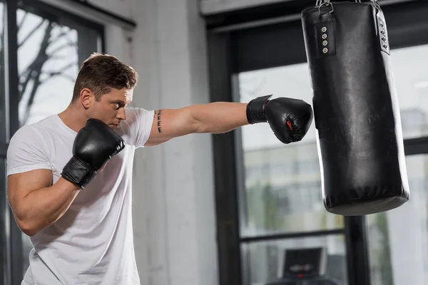 Vista lateral de belo boxeador muscular exercitando com saco de perfuração no ginásio — Fotografia de Stock