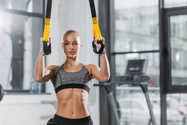 Attractive athletic sportswoman working out with suspension straps in gym and looking at camera — Stock Photo