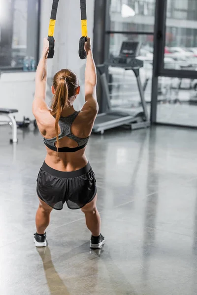 Back view of muscular sportswoman training with suspension straps in gym — Stock Photo