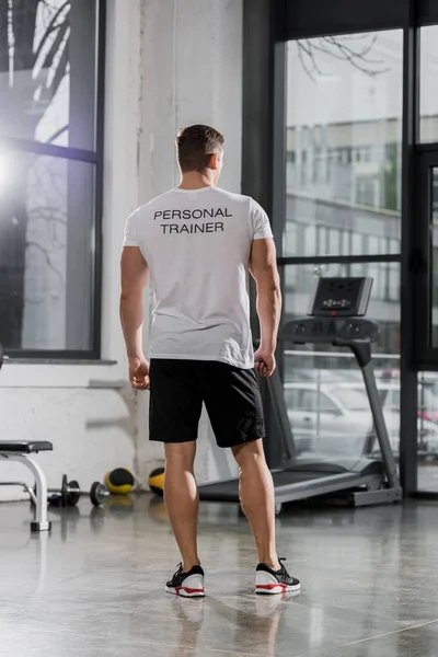 Vue arrière du bodybuilder athlétique en chemise avec texte entraîneur personnel debout dans la salle de gym — Photo de stock