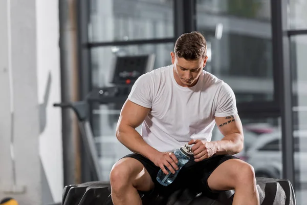 Beau bodybuilder athlétique assis sur le pneu avec bouteille d'eau sportive dans la salle de gym — Photo de stock