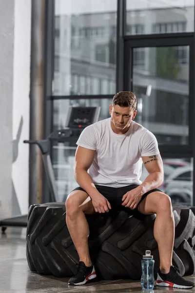 Fatigué beau bodybuilder athlétique assis sur le pneu dans la salle de gym — Photo de stock