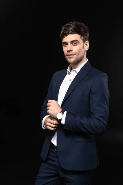 Joven hombre de negocios guapo en traje elegante con reloj de pulsera mirando a la cámara aislada en negro - foto de stock
