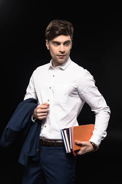 Handsome young businessman holding stack of notebooks isolated on black — Stock Photo