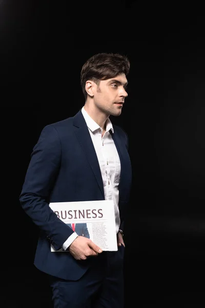 Attraktiver junger Geschäftsmann mit Zeitung, der isoliert auf schwarz schaut — Stockfoto