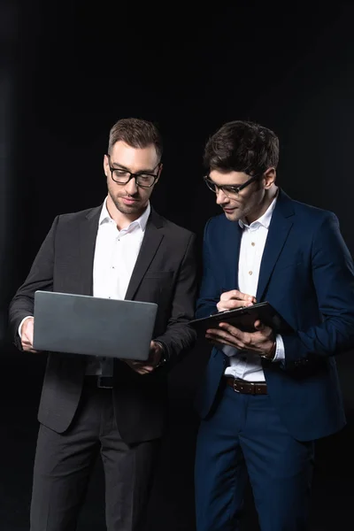 Впевнені молоді бізнесмени, що працюють з ноутбуком і буферами разом ізольовані на чорному — стокове фото