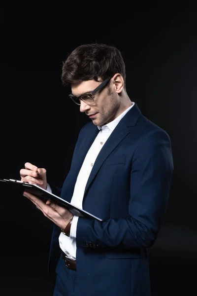 Handsome young businessman writing in clipboard isolated on black — Stock Photo