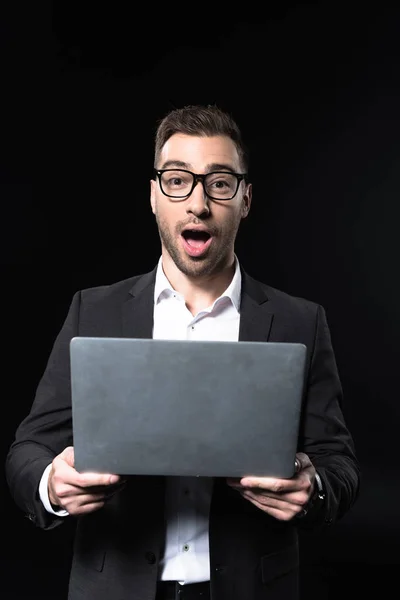 Емоційний молодий бізнесмен з ноутбуком дивиться на камеру ізольовано на чорному — стокове фото