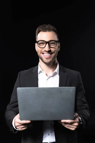 Smiling young businessman with laptop looking at camera isolated on black — Stock Photo