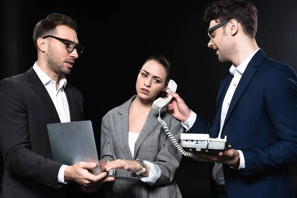 Overworked businesswoman talking by phone and working with laptop while colleagues supporting her isolated on black — Stock Photo