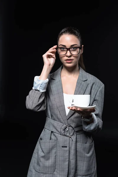 Elegante joven empresaria en chaqueta a cuadros sosteniendo taza de café y mirando a la cámara aislada en negro - foto de stock