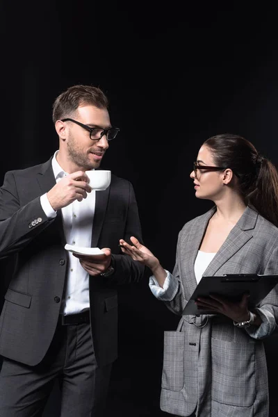 Business people with cup of coffee and clipboard having conversation isolated on black — Stock Photo