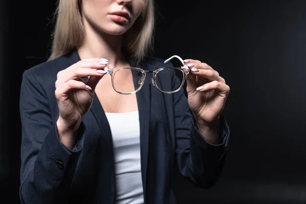 Plan recadré de jeune femme d'affaires en costume élégant tenant des lunettes isolées sur noir — Photo de stock