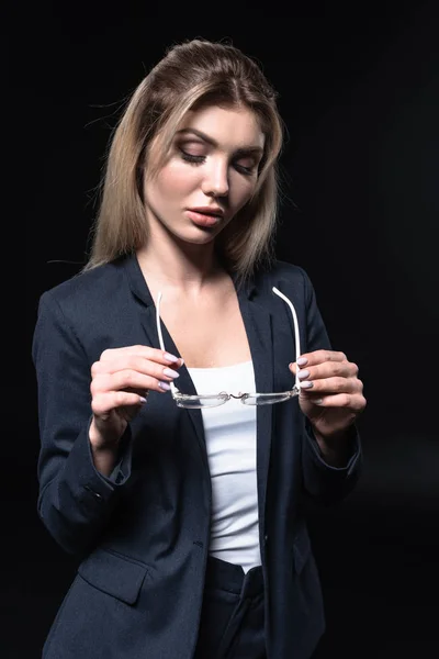 Attrayant jeune femme d'affaires en costume élégant tenant des lunettes isolées sur noir — Photo de stock