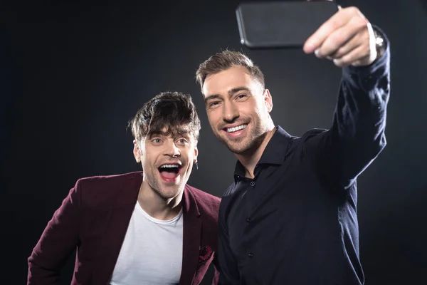 Jóvenes guapos en ropa elegante tomar selfie con teléfono inteligente en negro - foto de stock