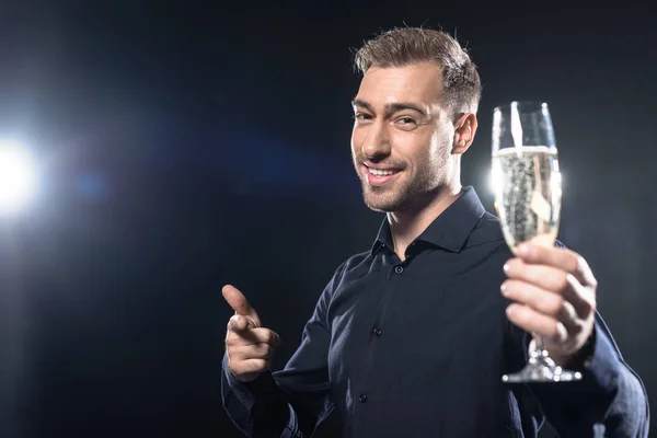 Lächelnder junger Mann im schicken Blazer mit Champagnerglas in der Hand und auf die Kamera gerichtet, isoliert auf schwarz — Stockfoto