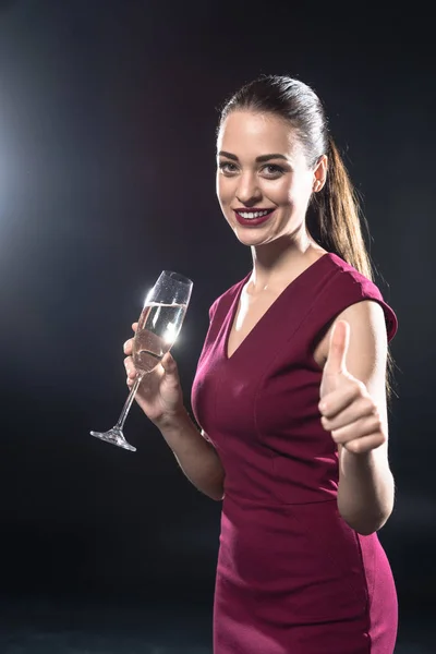 Lächelnde junge Frau hält ein Glas Champagner in der Hand und zeigt auf schwarz den Daumen in die Kamera — Stockfoto