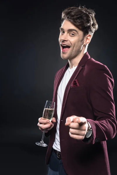 Felice giovane uomo in blazer elegante tenendo bicchiere di champagne e indicando la fotocamera isolata sul nero — Foto stock