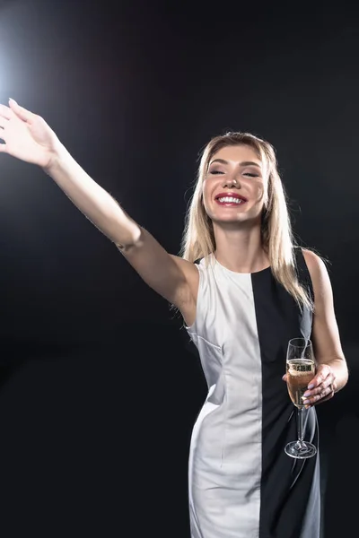 Feliz joven mujer sosteniendo la copa de champán y saludando con la mano en negro - foto de stock