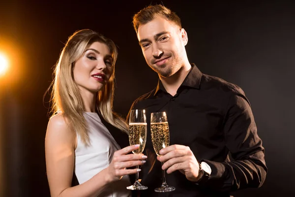 Heureux jeune couple avec des verres de champagne debout ensemble sur noir et regardant caméra — Photo de stock