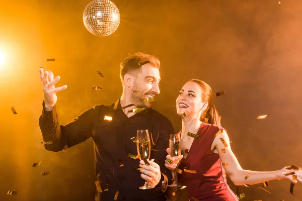 Glücklich flirtendes Paar mit Champagner, das sich bei einer Party unter goldenem Licht amüsiert, während Konfetti umfällt — Stockfoto