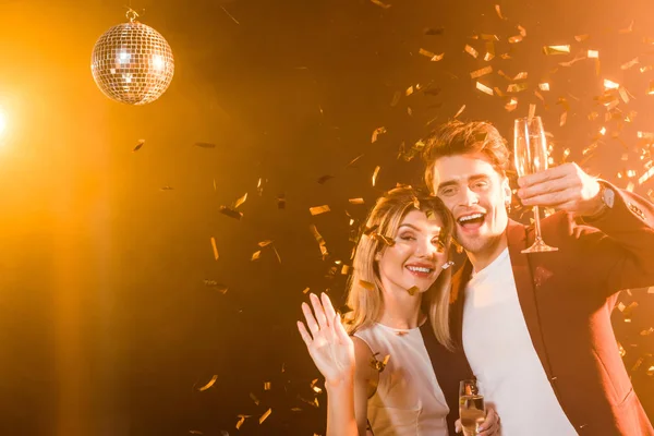 Happy flirting couple with champagne having fun during party under golden light while confetti spilling from above — Stock Photo