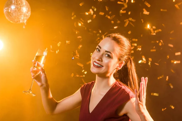 Emotional young woman with champagne glass under falling confetti, christmas concept — Stock Photo